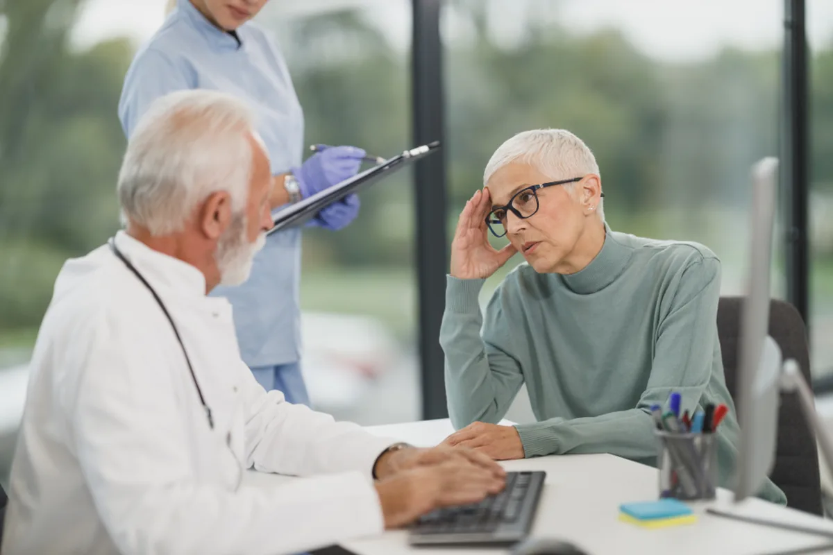 Physician at a computer reviewing a prior authorization request with a patient while a nurse holds a clipboard